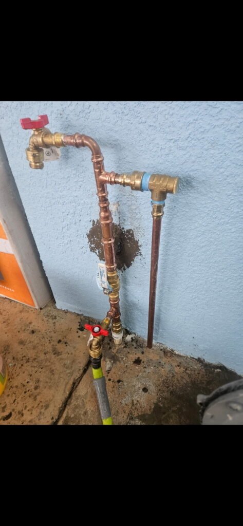 Exterior copper water piping with shutoff valves and hose connections against a blue wall.